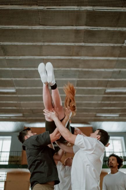 In a spacious indoor setting with a high, wooden-panel ceiling and large windows allowing natural light, two individuals are engaged in a team lifting technique to safely raise a young girl, who is we