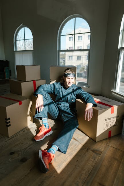 A man standing indoors in front of a plain, textured blue wall, surrounded by multiple cardboard boxes stacked on the wooden floor. The boxes vary in size and are sealed with red and black packing tap